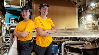 two female Mondi mill workers stand back to back with arms folded in a factory setting whilst wearing yellow t-shirts and black caps