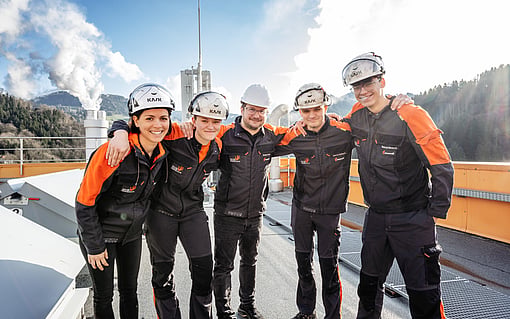 Five workers wearing protective helmets and orange‑and‑black uniforms stand together on an industrial site with mountains and steam in the background. They are smiling and have their arms around each other.