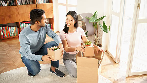 a man and a woman looking at each other in a home unboxing house plants with a bookshelf behind them and a rug beneath them