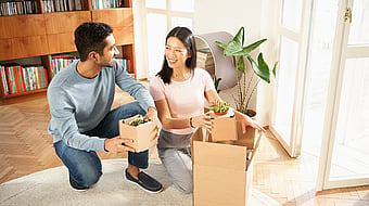 a man and a woman looking at each other in a home unboxing house plants with a bookshelf behind them and a rug beneath them