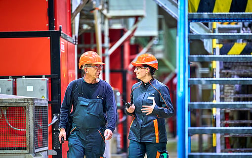 Two workers wearing orange safety helmets and dark protective uniforms walk together inside an industrial facility, engaged in conversation. They are surrounded by large machinery, metal structurer and bright industrial equipment.