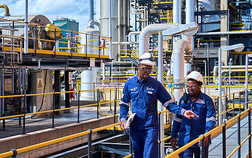 two workers in blue overalls and white hardhats talking with a factory background