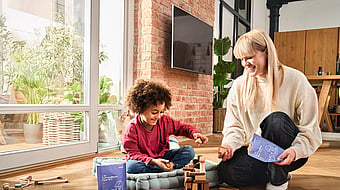 A woman and a child sitting together by a window while playing, with the woman holding a Mondi package.