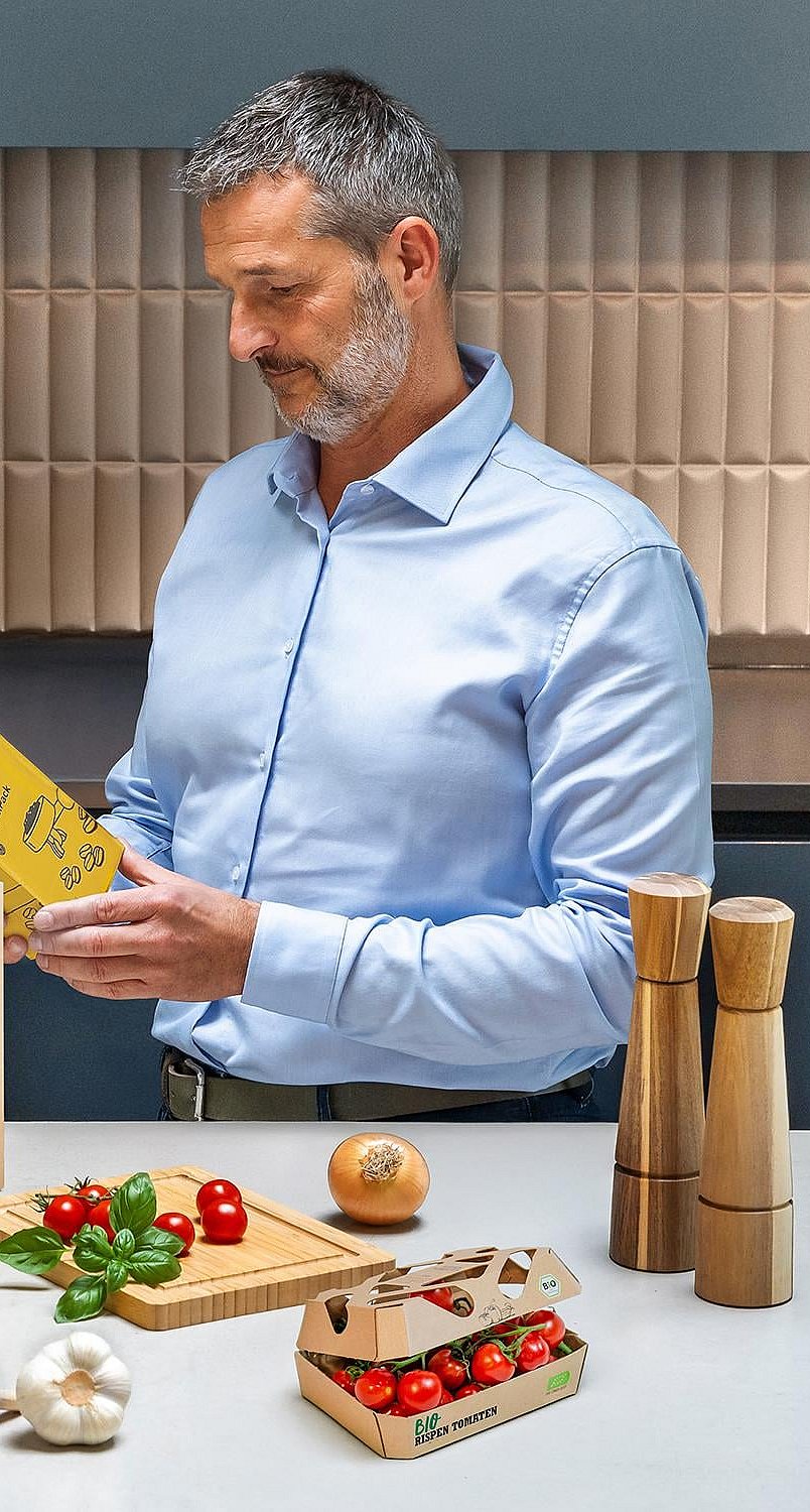 man standing at a kitchen counter reading the text on some food packaging