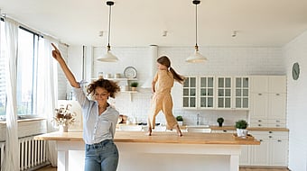 Woman dancing in a bright white kitchen while a child dances on top of the kitchen island, with large windows letting in natural light