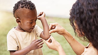 young child in beige t-shirt getting a plaster stuck on his left arm by a woman