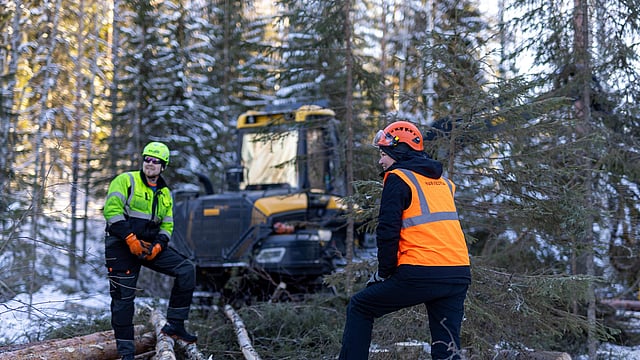 Two forestry workers wearing high‑visibility safety gear stand among felled logs in a snowy forest, with a large forestry machine in the background.