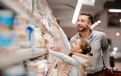 A smiling man is standing in front of a cooling shelf at a supermarket with a child.