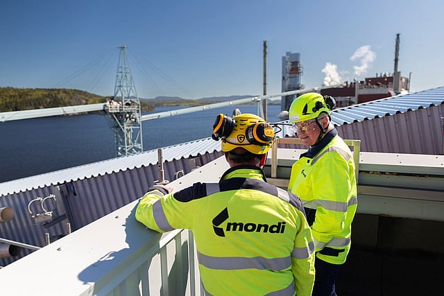 Two Mondi workers in high‑visibility safety gear stand on an industrial rooftop overlooking a waterfront facility with cranes, piping, and processing buildings in the background.