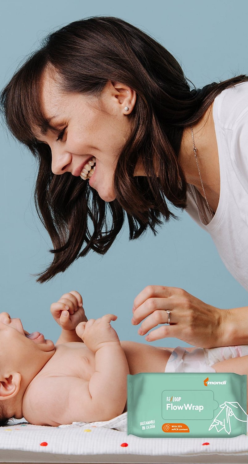 Person beugt sich über ein Baby, das auf einer Wickelunterlage liegt. Im Vordergrund ist eine Packung Feuchttücher für Babys zu sehen. Die Szene zeigt einen Betreuungsmoment in einem Innenraum vor einem hellblauen Hintergrund mit weicher Beleuchtung.