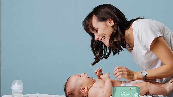 Person leaning over a baby lying on a changing mat in an indoor setting. A pack of baby wipes is visible in the foreground. The scene focuses on a caregiving moment between the person and baby, with a light blue background and soft natural lighting.