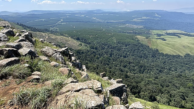 A panoramic view from a rocky hillside overlooking a vast landscape of forests, fields, and distant rolling hills under a clear blue sky.