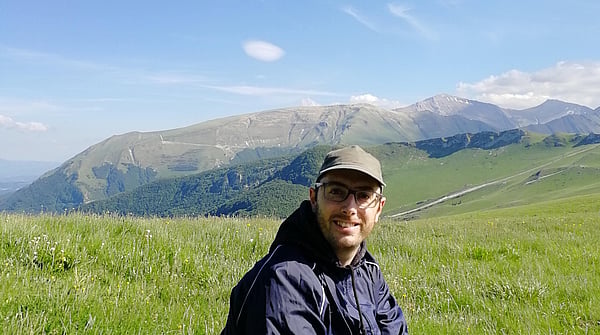 man in cap and hooded jacket sat on a hillside with a blue sky and hills in the background