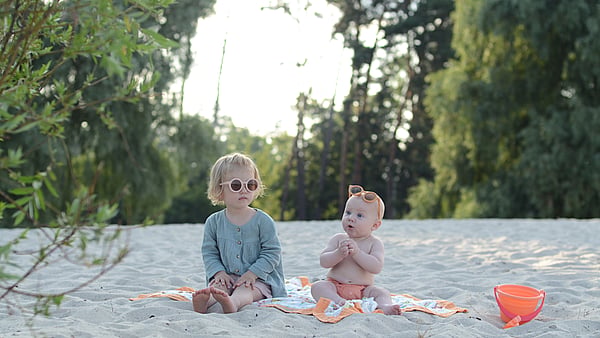 Two toddlers sit next to each other on a beach wearing orange sunglasses with beach toys nearby.