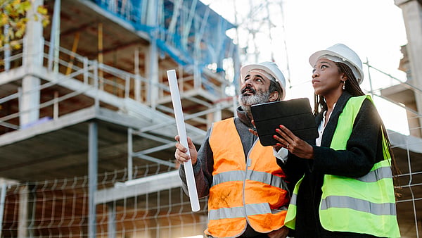 a man and a lady in safety gear stood looking up on a building site while holding a clipboard