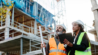 a man and a lady in safety gear stood looking up on a building site while holding a clipboard
