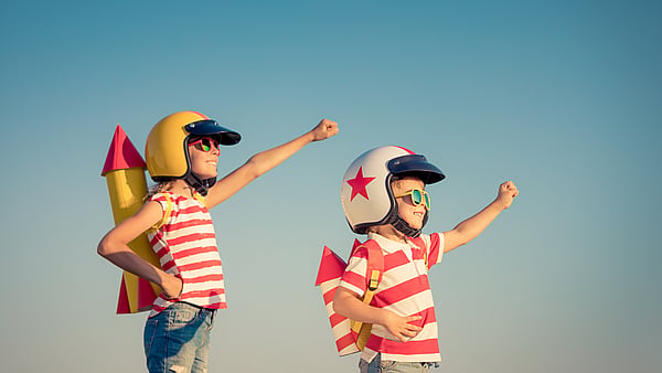 two children both wearing red and white stripey t-shirts and sunglasses dressed up as spacemen with their fists raised set against a blue sky