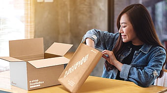 woman reaching into a package as she sits at a table with another cardboard box nearby