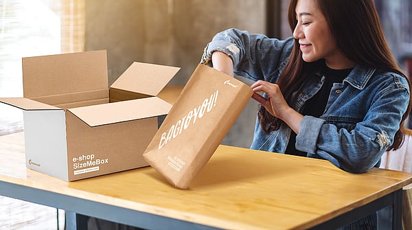 woman reaching into a package as she sits at a table with another cardboard box nearby