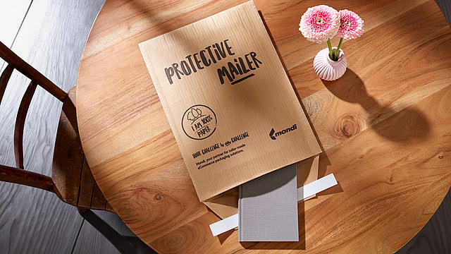 A Protective Mailer is open on a table, with a book half-packed into it and a vase with a flower next to it.