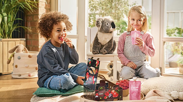 Two children sitting next to a koala toy, smiling and holding a drinking pouch.
