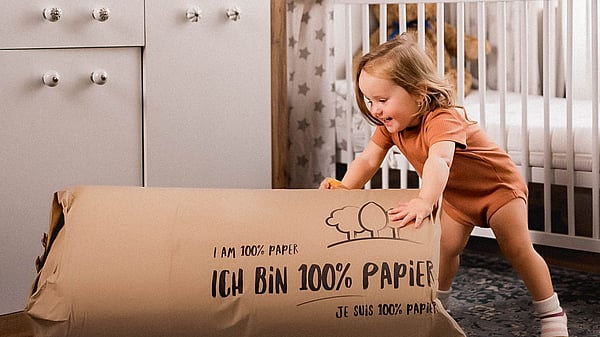young girl in orange t-shirt and orange shorts leaning on a large piece of cardboard packaging and smiling with a crib behind her