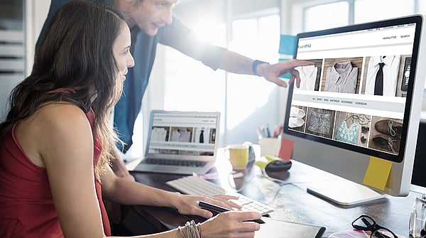 a lady sat at a desktop computer in an office while a man stands beside her pointing at the screen