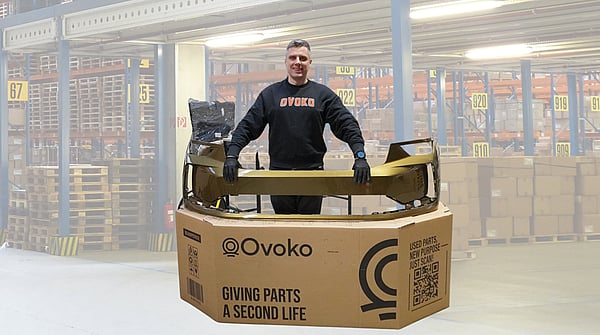 Person standing behind a large cardboard shipping box while holding a gold-coloured automotive bumper part placed across the top. The box is printed with the “Ovoko” logo and the text “Giving parts a second life,” along with a QR code and handling icons. The person is wearing dark clothing and gloves. Additional packaging material and a wrapped item are visible behind the box, all set against a plain white background.