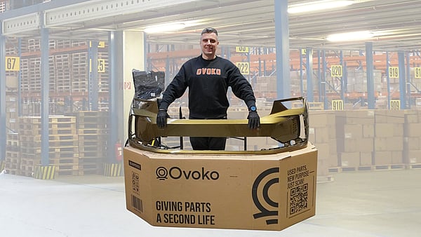 Person standing behind a large cardboard shipping box while holding a gold-coloured automotive bumper part placed across the top. The box is printed with the “Ovoko” logo and the text “Giving parts a second life,” along with a QR code and handling icons. The person is wearing dark clothing and gloves. Additional packaging material and a wrapped item are visible behind the box, all set against a plain white background.