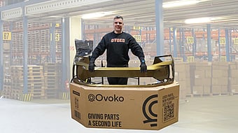 Person standing behind a large cardboard shipping box while holding a gold-coloured automotive bumper part placed across the top. The box is printed with the “Ovoko” logo and the text “Giving parts a second life,” along with a QR code and handling icons. The person is wearing dark clothing and gloves. Additional packaging material and a wrapped item are visible behind the box, all set against a plain white background.