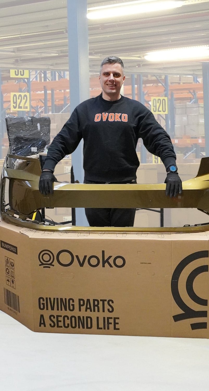 Person standing behind a large cardboard shipping box while holding a gold-coloured automotive bumper part placed across the top. The box is printed with the “Ovoko” logo and the text “Giving parts a second life,” along with a QR code and handling icons. The person is wearing dark clothing and gloves. Additional packaging material and a wrapped item are visible behind the box, all set against a plain white background.