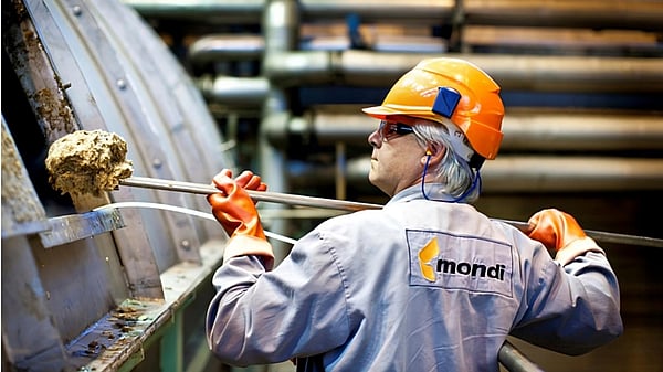 A Mondi employee wearing grey overalls, safety goggles and an orange hardhat picks up residues from wood fibres, chemicals and other substances from a large machine.