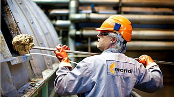 A Mondi employee wearing grey overalls, safety goggles and an orange hardhat picks up residues from wood fibres, chemicals and other substances from a large machine.