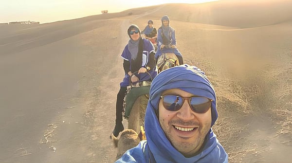 Image of Youssef Agzenai wearing a blue headscarf and sunglasses in a desert riding a camel followed by young children also riding a camel