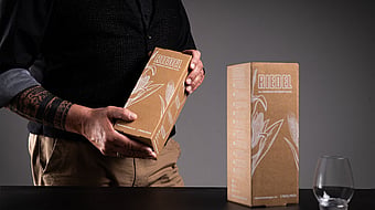man in black clothing holding a brown corrugated box with a glass on the table next to him