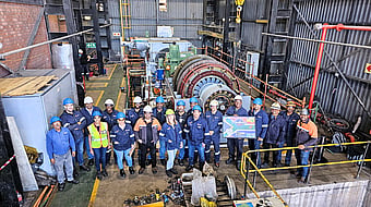 large group of people pose in front of factory machinery wearing hi-viz and blue jackets and hard hats