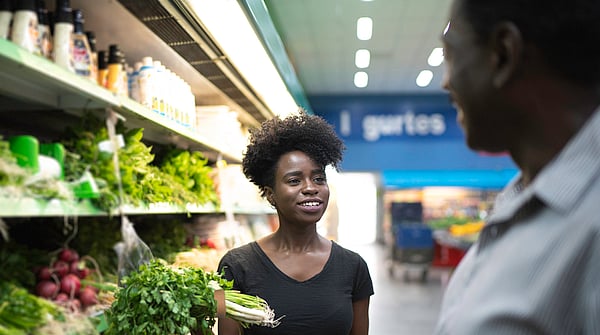 lady wearing a black top holding fresh vegetables in front of shelves of other fresh vegetables whilst talking to a man