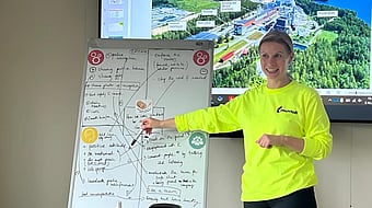 woman in bright jumper standing in front of a screen and a whiteboard whilst addressing an audience