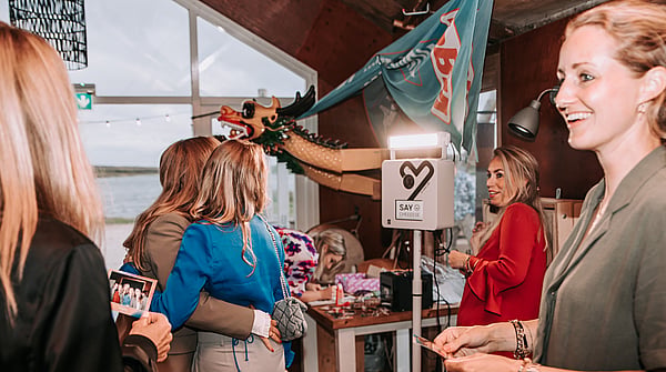 Two women are hugging and taking a picture with the KRUU photo booth. There are also two other women in the picture.