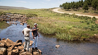 two men in neutral clothing on the edge of a river with a forest in the background. one of the men is pointing towards the forest